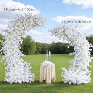 May include: Two white floral arches, one 100 inches and the other 80 inches, stand on a grassy field. A small round table with a lace tablecloth and candles is between the arches. The sky is blue with scattered clouds.