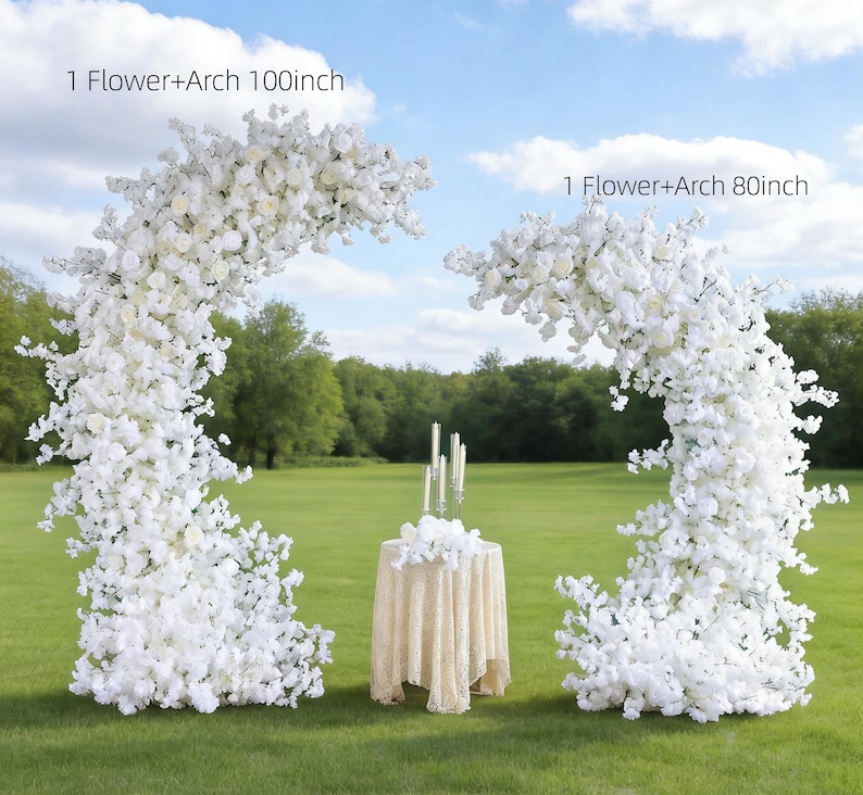 May include: Two white floral arches, one 100 inches and the other 80 inches, stand on a grassy field. A small round table with a lace tablecloth and candles is between the arches. The sky is blue with scattered clouds.
