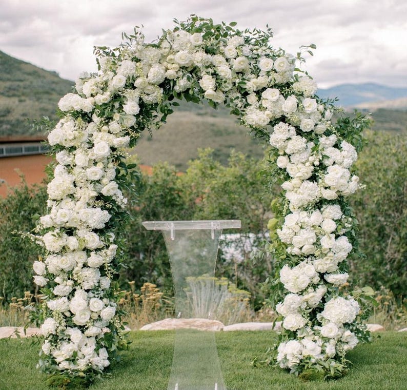 May include: A white floral archway made of white flowers and greenery, with a clear acrylic podium in front of it.
