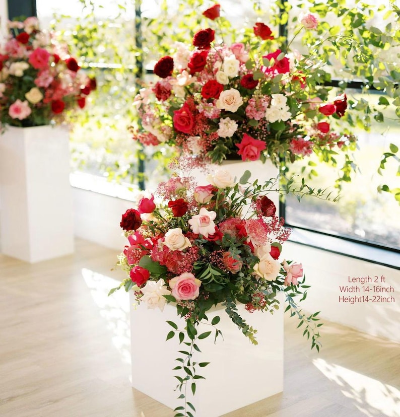 May include: Three white square pedestals each hold a vibrant floral arrangement. The flowers are a mix of red, pink, and white roses, with greenery. The text on the image reads: Length 2 ft, Width 14-16 inch, Height 14-22 inch.