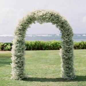 May include: A white flower archway made of baby's breath flowers. The archway is set up outdoors on a grassy lawn with a view of the ocean in the background.