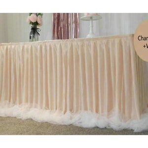 May include: Champagne and white table skirt with a white tulle ruffle. The skirt is draped over a table with a vase of pink flowers and a cake stand on top.