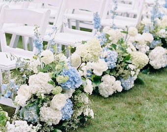 Puede incluir: Arreglos florales de hortensias blancas y azules, rosas y otras flores bordean una fila de sillas plegables blancas sobre césped. Los arreglos miden aproximadamente 61 cm de largo, 36-41 cm de ancho y 41-61 cm de alto.