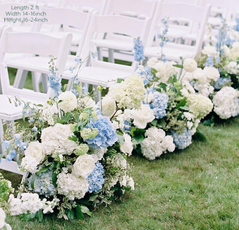 May include: White folding chairs line a grassy aisle, decorated with floral arrangements. The arrangements include white roses, hydrangeas, and blue delphiniums. The text in the image reads: Length 61 cm, Width 36-41 cm, Height 41-61 cm.