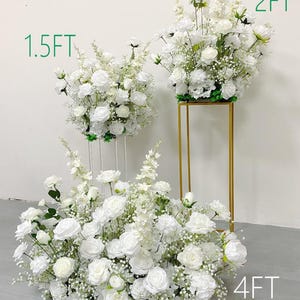 May include: Arrangement of white roses and other white flowers. The arrangement is 4 feet wide, with other arrangements at 1.5 feet and 2 feet. The flowers are on a gold stand and clear stands.