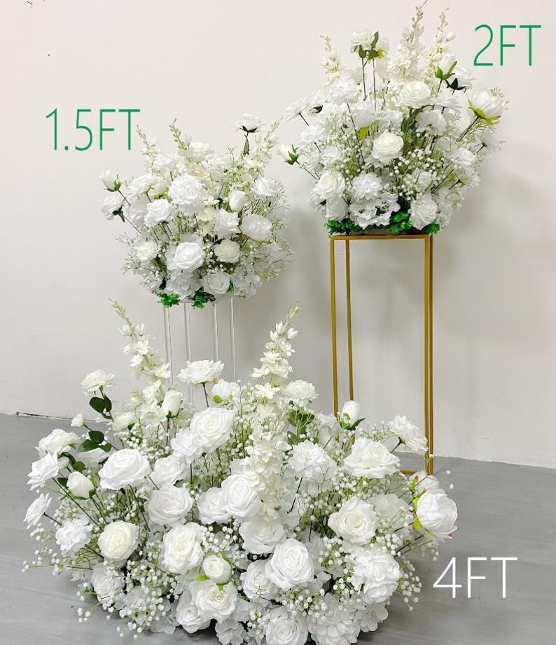 May include: Arrangement of white roses and other white flowers. The arrangement is 4 feet wide, with other arrangements at 1.5 feet and 2 feet. The flowers are on a gold stand and clear stands.