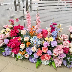 May include: A large, dense floral arrangement featuring a vibrant mix of artificial flowers in shades of pink, purple, blue, orange, and cream. The display includes roses, hydrangeas, and tall spikes of pink blossoms, accented with green leaves.