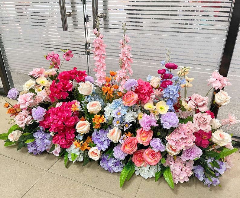 May include: A large, dense floral arrangement featuring a vibrant mix of artificial flowers in shades of pink, purple, blue, orange, and cream. The display includes roses, hydrangeas, and tall spikes of pink blossoms, accented with green leaves.
