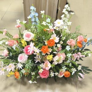 May include: A colourful floral display with pink roses, orange ranunculus, and white flowers. The arrangement is set against a backdrop of green foliage. The flowers are a mix of textures and shades, creating a visually appealing display.