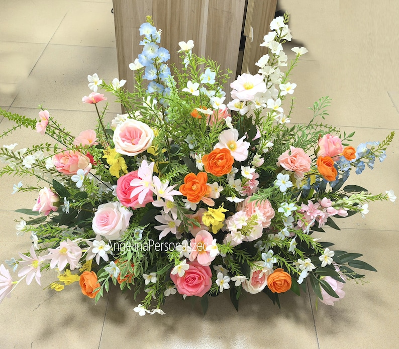 May include: A colourful floral display with pink roses, orange ranunculus, and white flowers. The arrangement is set against a backdrop of green foliage. The flowers are a mix of textures and shades, creating a visually appealing display.