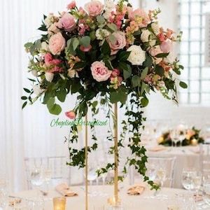 May include: A tall gold metal centerpiece with a large arrangement of pink and white roses, greenery, and other flowers. The centerpiece is on a white tablecloth with clear glass glasses and candles.
