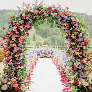 May include: A wedding ceremony archway decorated with a lush floral arrangement of pink, red, and blue flowers. The archway is set up outdoors with a white aisle runner and pink flower petals scattered on the ground.