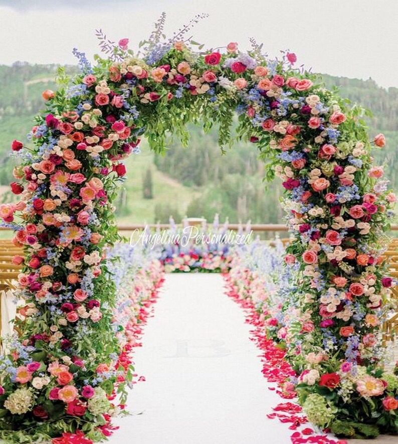 May include: A wedding ceremony archway decorated with a lush floral arrangement of pink, red, and blue flowers. The archway is set up outdoors with a white aisle runner and pink flower petals scattered on the ground.