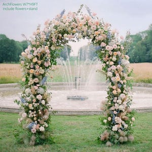 K&ouml;nnte beinhalten: Ein Blumenbogen, ca. 213 cm breit und 274 cm hoch, geschm&uuml;ckt mit pastellfarbenen Blumen und Gr&uuml;n. Der Bogen steht im Freien auf einer Wiese, mit einem Brunnen im Hintergrund. Der Text "ArchFlowers W7*H9FT include flowers+stand" ist sichtbar.