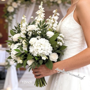 Puede incluir: Un ramo de novia con rosas blancas, hortensias y otras flores blancas con follaje verde. El ramo mide aproximadamente 30 cm de largo y lo sostiene una persona con un vestido de novia blanco.