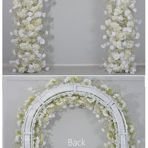 K&ouml;nnte beinhalten: Ein wei&szlig;er Metallbogenst&auml;nder, verziert mit wei&szlig;en Blumen und Gr&uuml;n. Der Bogen ist mit wei&szlig;en Blumen und Gr&uuml;n bedeckt und bildet eine sch&ouml;ne Kulisse f&uuml;r Hochzeiten oder Veranstaltungen. Der Text "Back arch stand" ist sichtbar.
