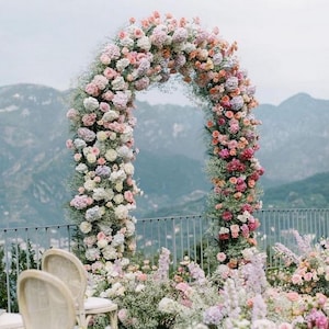 May include: A floral archway made of pink, white, and purple flowers, with a backdrop of mountains and a blue sky. The archway is decorated with greenery and baby's breath.