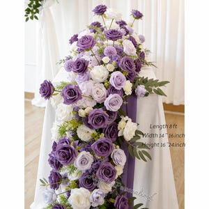 May include: A floral arrangement featuring purple and white roses, lavender hydrangeas, and greenery. The arrangement is displayed on a white tablecloth with a purple runner. The text on the image reads: Length 8 ft, Width 14-16 inch, Height 16-24 inch.