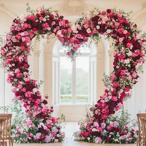 May include: A heart-shaped floral arch made of pink and burgundy flowers, greenery, and leaves. The arch is in front of a large window with a view of trees.