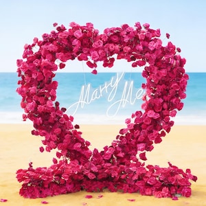 May include: A large heart-shaped floral arrangement made of vibrant pink roses. The heart frames the words "Marry Me" in elegant white script, set against a beach backdrop with a blue sky and ocean.