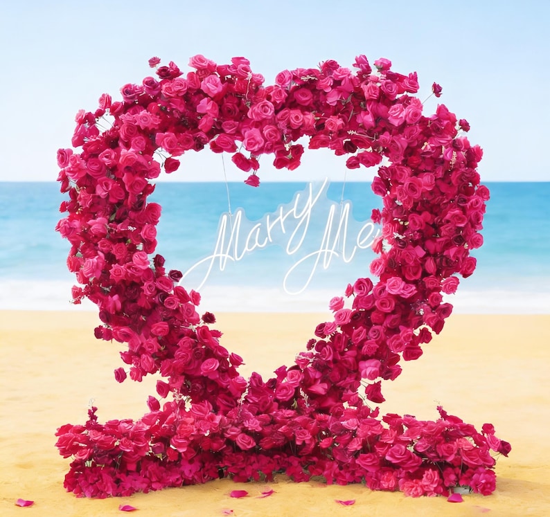 May include: A large heart-shaped floral arrangement made of vibrant pink roses. The heart frames the words "Marry Me" in elegant white script, set against a beach backdrop with a blue sky and ocean.
