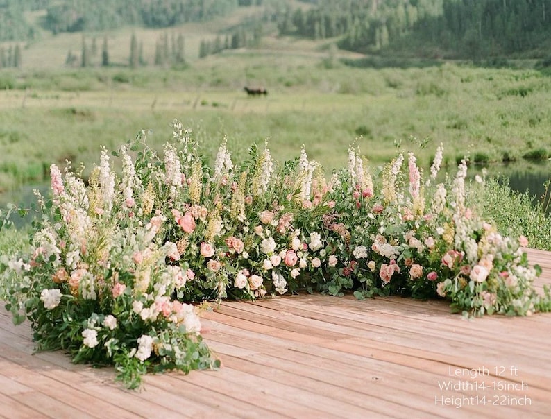 May include: A floral archway with a wooden base, decorated with pink, white, and yellow flowers. The arch is set against a green field and water. The text on the image says: Length 3.66 m, Width 35.56-40.64 cm, Height 35.56-55.88 cm.