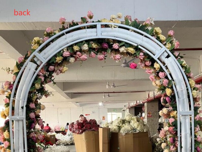 May include: White metal archway decorated with pink, white, and yellow artificial flowers. The archway is in a warehouse setting.