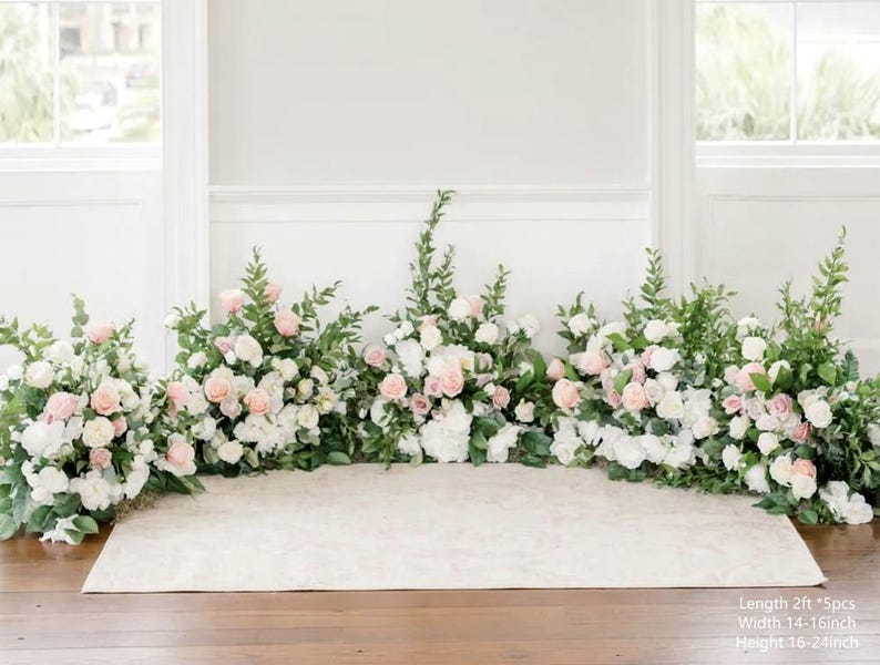 Op de afbeelding: Een witte achtergrond versierd met een bloemstuk van witte en roze bloemen en groen. De bloemen zijn in een lange lijn over de achtergrond gerangschikt, wat een romantische en elegante uitstraling cre&euml;ert. De achtergrond is perfect voor een huwelijksceremonie of andere speciale gelegenheid.