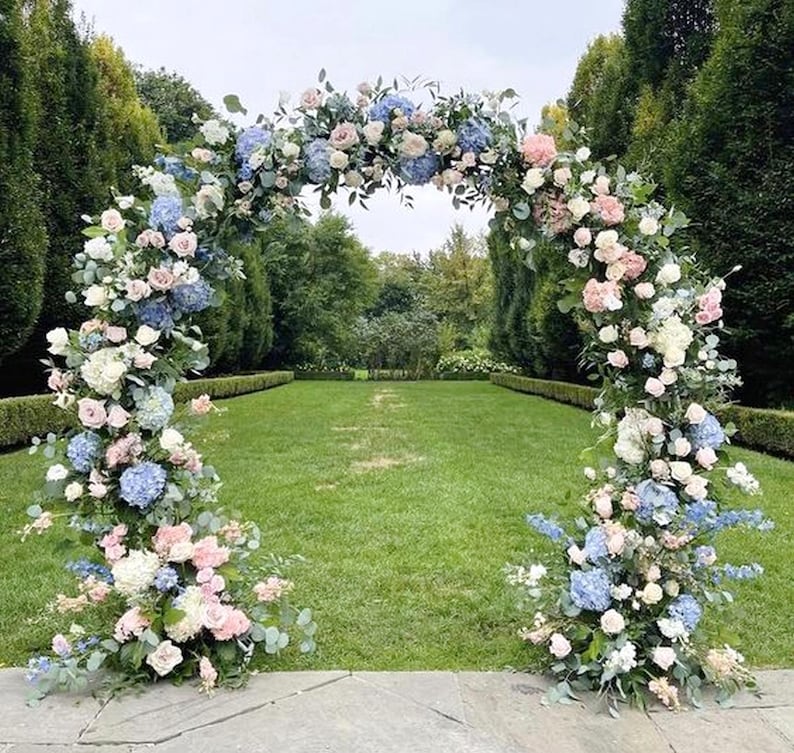 May include: A wedding arch decorated with blue and pink flowers, including hydrangeas and roses. The arch is set up outdoors in a grassy area.
