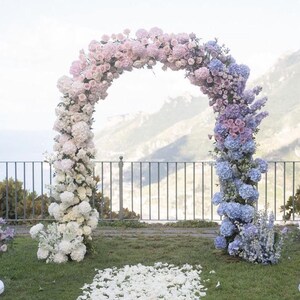 May include: A wedding arch decorated with a gradient of flowers, starting with white and transitioning to pink and then blue. The arch is outdoors with a view of a mountain range in the background.