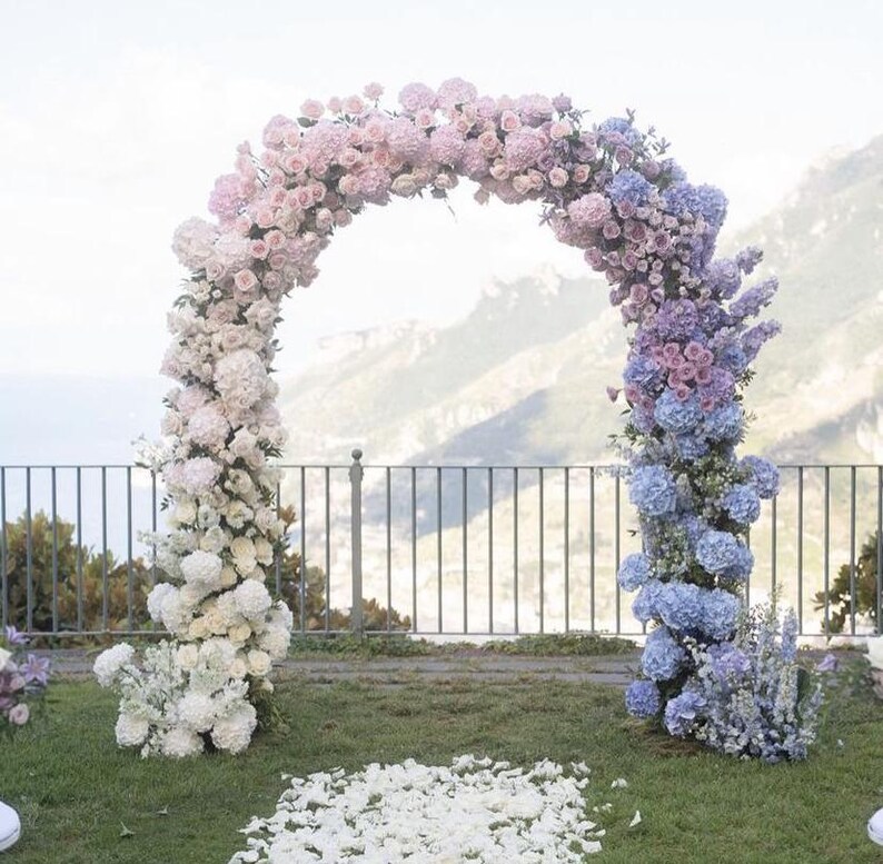 May include: A wedding arch decorated with a gradient of flowers, starting with white and transitioning to pink and then blue. The arch is outdoors with a view of a mountain range in the background.