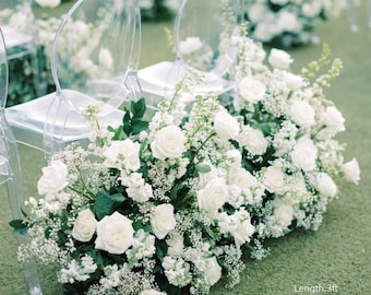 Op de afbeelding: Witte rozen bloemstukken met groen aan de basis van doorzichtige stoelen. De arrangementen zijn 91 cm lang, 36-41 cm breed en 41-56 cm hoog. De tekst in de afbeelding luidt: Length 3ft, Width 14-16inch, Height 16-22inch.