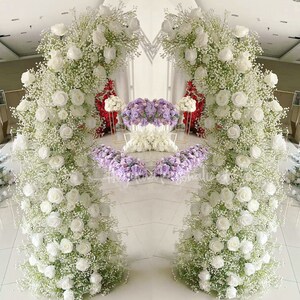 May include: A wedding arch decorated with white flowers and greenery. The arch is made of two separate pieces, each with a white flower arrangement. The arch is set up in a room with a white floor and white walls.
