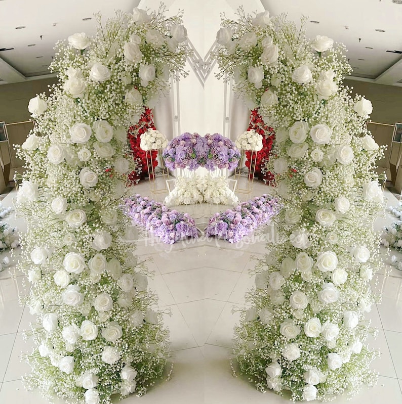 May include: A wedding arch decorated with white flowers and greenery. The arch is made of two separate pieces, each with a white flower arrangement. The arch is set up in a room with a white floor and white walls.