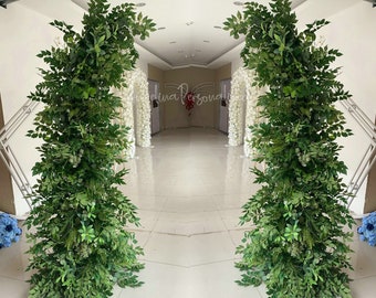 May include: Two tall green leafy archways made of artificial greenery, with white flower arrangements behind them, stand in a hallway with a tiled floor.