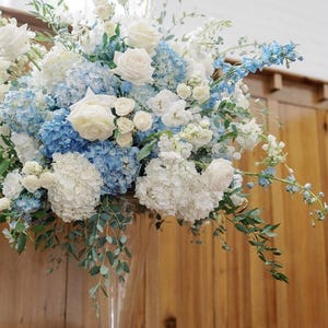 Op de afbeelding: Een groot bloemstuk met witte rozen, blauwe hortensia's en andere witte en blauwe bloemen. Het arrangement wordt gepresenteerd in een heldere glazen vaas. De breedte van het arrangement is 56 cm.