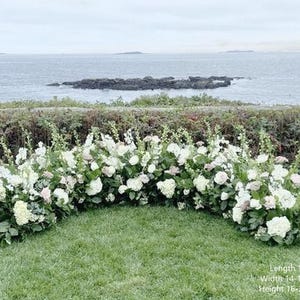 Op de afbeelding: Een witte bloemenboog met groen, perfect voor een huwelijksceremonie of andere speciale gelegenheid. De boog is 3 meter lang, 35-40 cm breed en 40-60 cm hoog.