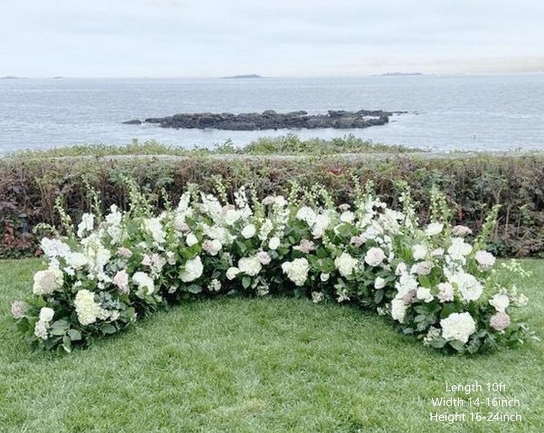 Op de afbeelding: Een witte bloemenboog met groen, perfect voor een huwelijksceremonie of andere speciale gelegenheid. De boog is 3 meter lang, 35-40 cm breed en 40-60 cm hoog.