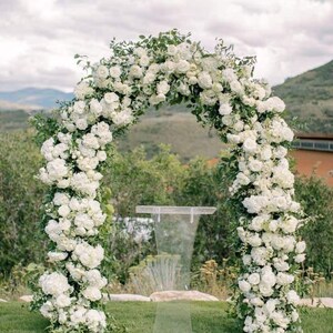 May include: A wedding arch adorned with lush white flowers and greenery, creating a romantic outdoor setting. A clear acrylic podium stands in front of the arch, set on a green lawn with a backdrop of rolling hills and a cloudy sky.