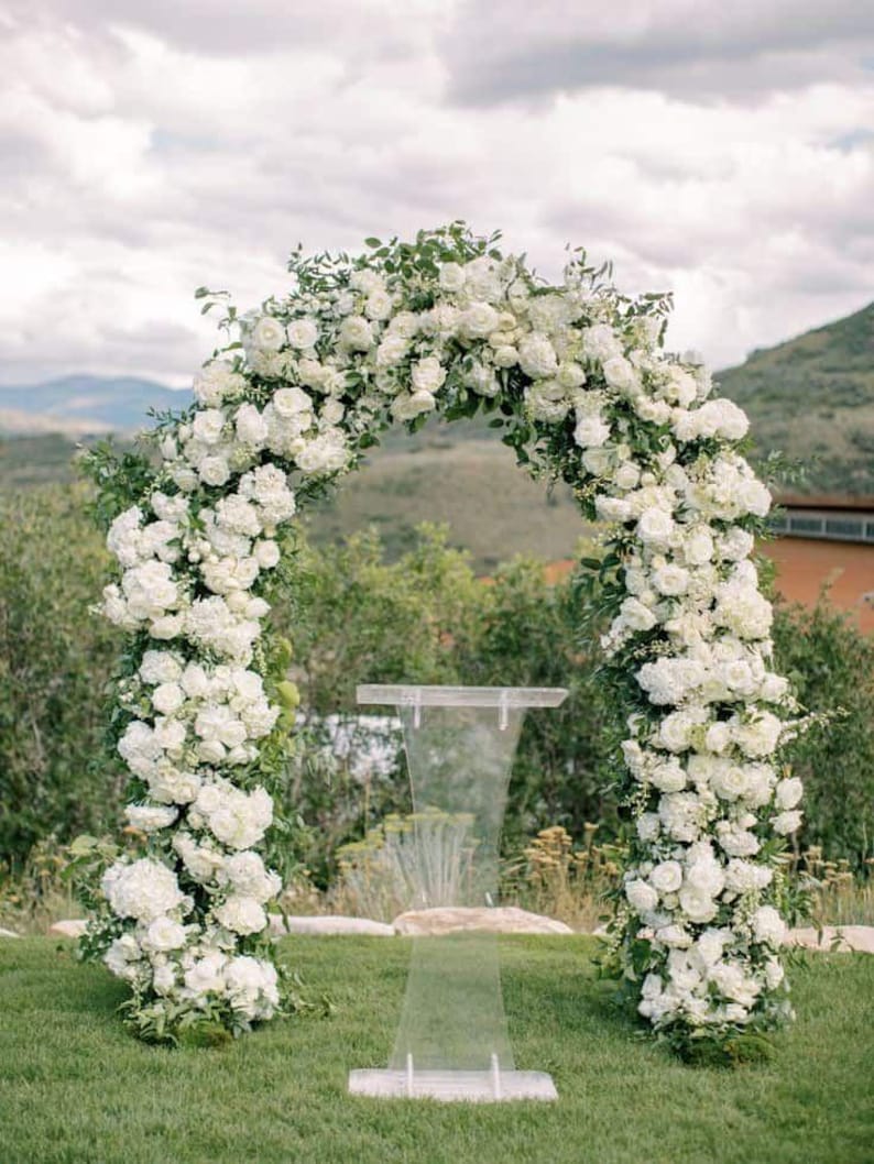 May include: A wedding arch adorned with lush white flowers and greenery, creating a romantic outdoor setting. A clear acrylic podium stands in front of the arch, set on a green lawn with a backdrop of rolling hills and a cloudy sky.
