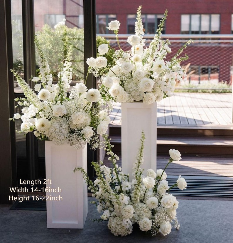 May include: Two white rectangular pedestals of varying heights, each topped with a large arrangement of white roses, hydrangeas, and tall snapdragons. A third floral arrangement sits on the ground. Text states Length 2ft, Width 14-16inch, Height 16-22inch.