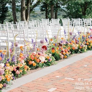 Puede incluir: Un pasillo de ceremonia de boda decorado con un arreglo floral colorido de flores naranjas, amarillas, moradas y rojas. Las flores est&aacute;n dispuestas en una fila a lo largo del pasillo, que est&aacute; hecho de ladrillo. Las sillas blancas est&aacute;n dispuestas en filas a ambos lados del pasillo.