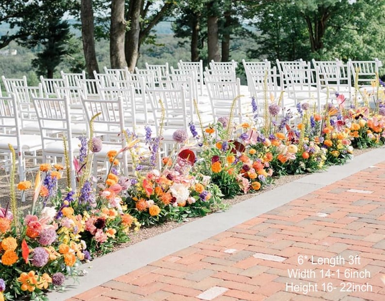 Puede incluir: Un pasillo de ceremonia de boda decorado con un arreglo floral colorido de flores naranjas, amarillas, moradas y rojas. Las flores est&aacute;n dispuestas en una fila a lo largo del pasillo, que est&aacute; hecho de ladrillo. Las sillas blancas est&aacute;n dispuestas en filas a ambos lados del pasillo.