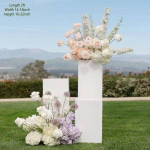 May include: White rectangular pedestals with floral arrangements. The pedestals are 61 cm long, 36-41 cm wide, and 41-56 cm high. The flowers include roses and hydrangeas in shades of white, pink, and purple, set against a green lawn and a blue sky.