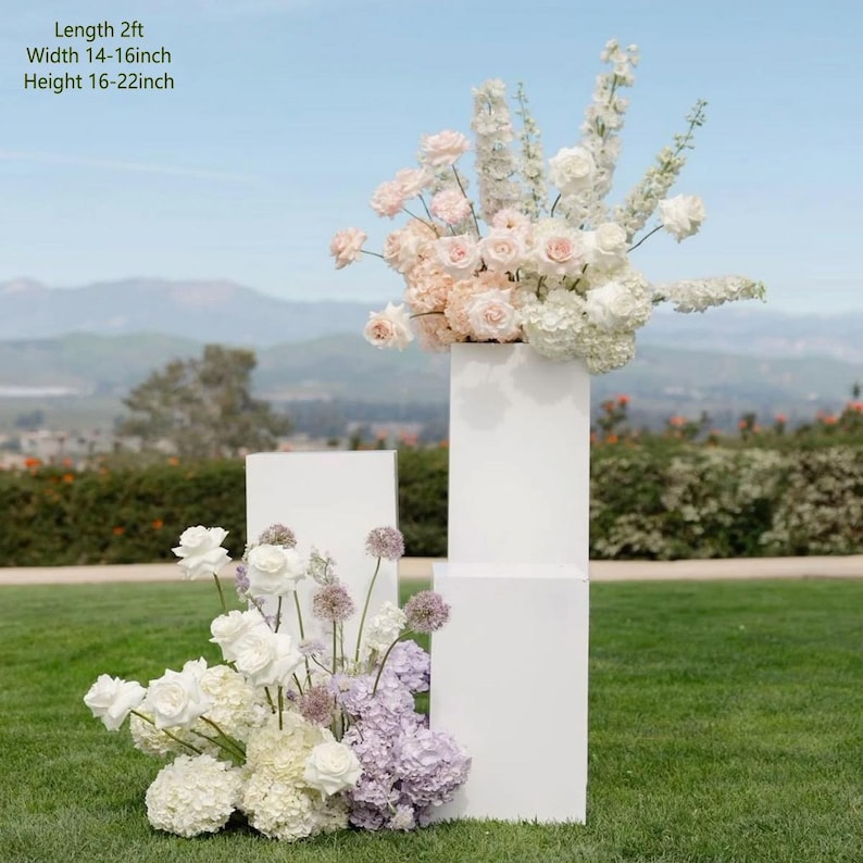 May include: White rectangular pedestals with floral arrangements. The pedestals are 61 cm long, 36-41 cm wide, and 41-56 cm high. The flowers include roses and hydrangeas in shades of white, pink, and purple, set against a green lawn and a blue sky.