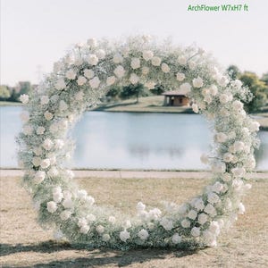 May include: A white floral arch made of baby's breath and white roses, measuring 7 feet wide by 7 feet high. The arch is set up outdoors in front of a lake.