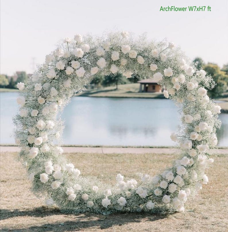 May include: A white floral arch made of baby's breath and white roses, measuring 7 feet wide by 7 feet high. The arch is set up outdoors in front of a lake.