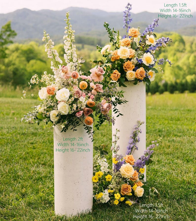 May include: Two white cylindrical pedestals with floral arrangements. The arrangements include roses, delphiniums, and other flowers in peach, yellow, and white. The pedestals are on grass with mountains behind. Length 61cm, Width 36-41cm, Height 41-56cm.