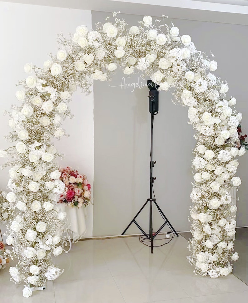 May include: A white floral archway made of white roses and baby's breath, perfect for a wedding ceremony or special event. The archway is standing in a room with a white wall and a black tripod stand.
