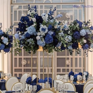 Puede incluir: Arreglos florales elegantes en tonos azules y blancos, exhibidos sobre soportes de metal dorado. Los arreglos miden 51 cm de ancho y están colocados sobre mesas con sillas blancas y ribetes dorados, creando un ambiente sofisticado.
